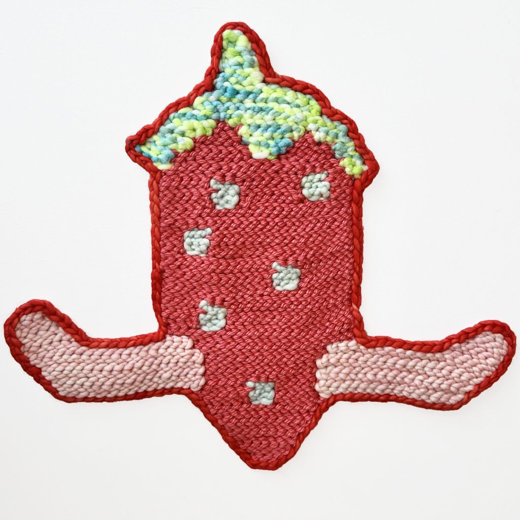 A handmade woven textile art piece shaped like a red strawberry with green and yellow at the top, featuring two pink, wing-like shapes extending from either side, against a white background.