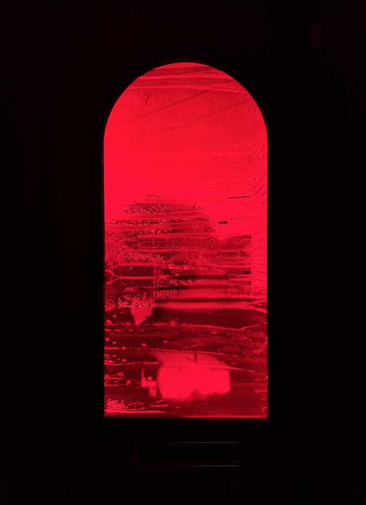 An arched window glows with intense red light from the inside, partially obscured by a photographic transfer. A distorted view of the outside world creates a feeling of enclosure and longing.
