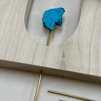 A bright blue stone rests on a gold metal rod, positioned in a cutout area of a light wood board. A red mechanical pencil lies nearby on a white surface.