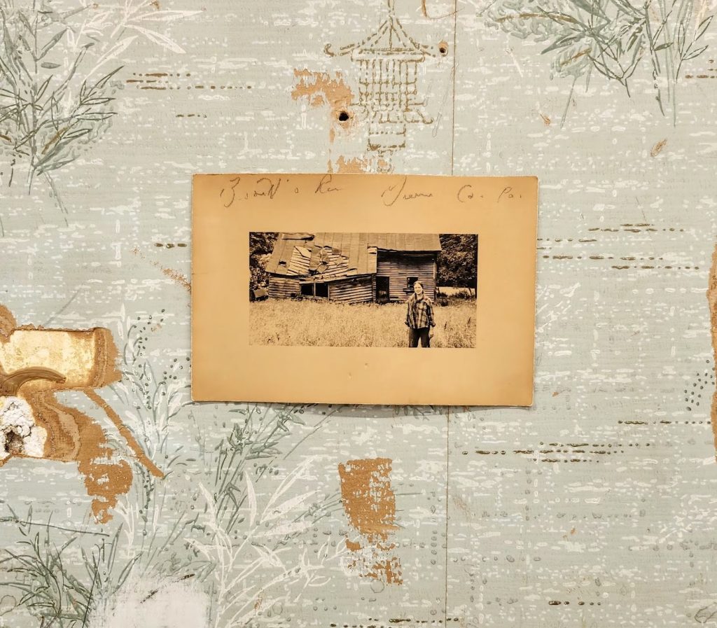 A sepia-toned photo of a person standing in front of a rustic wooden cabin is pinned to a faded, patterned wall with peeling wallpaper and handwritten text above the photo.