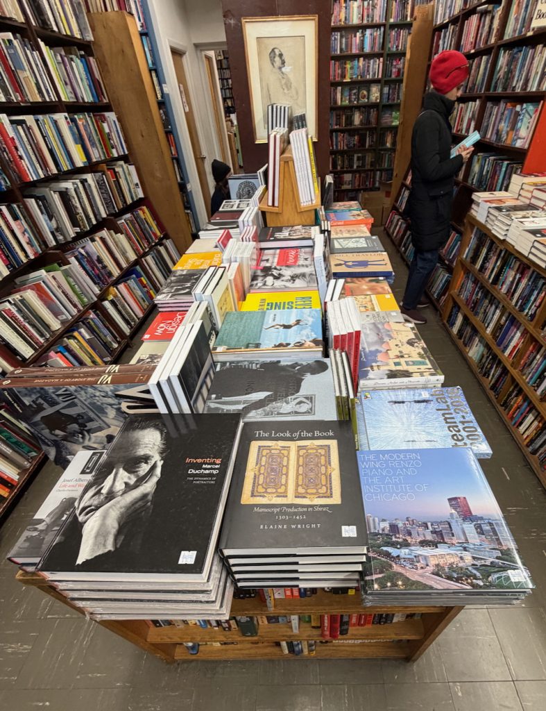 A table filled with neatly stacked books sits between tall shelves lined with more books in a bookstore; a person in a red hat browses the shelves in the background.