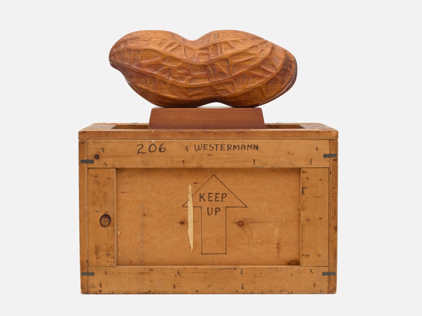A wooden sculpture shaped like a peanut sits atop a wooden crate labeled “KEEP UP” with an upward arrow, and the text “WESTERMANN” and “206” written on the crate. The background is plain white.