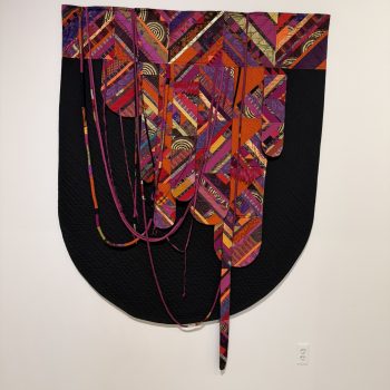 A textile artwork featuring vibrant, multicolored geometric patterns draped over a large, rounded black quilted background, with long fabric strips hanging down. The piece is displayed on a plain white wall.