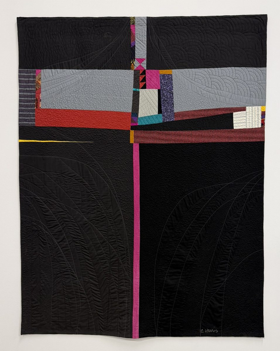 A modern quilt with abstract geometric shapes in black, gray, red, yellow, and magenta, featuring intersecting lines and textured stitching patterns, hangs against a plain white wall.