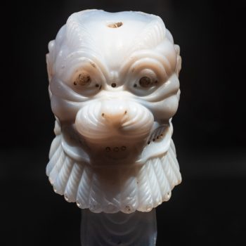 A close-up of a carved white jade sculpture resembling a stylized animal or mythical creatures head, with detailed facial features and textured mane, displayed on a black stand against a dark background.