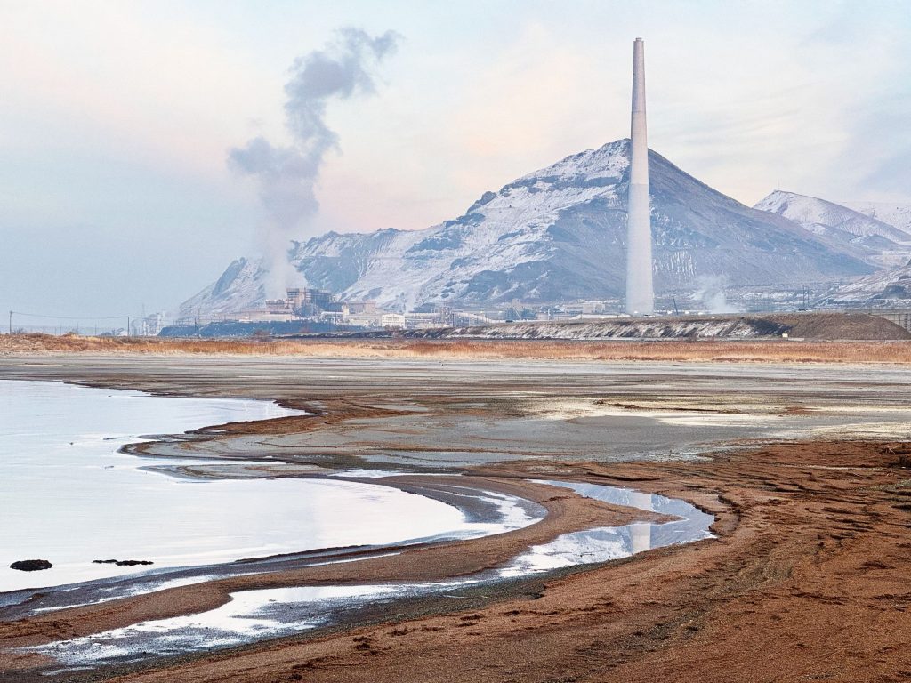 Industrial smokestack rises near snow-covered mountains, emitting smoke. A lake and sandy shore are in the foreground, under a pale sky.