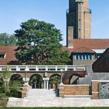 A tall brick observatory tower with a domed roof rises behind an arched brick walkway, mature trees, and a manicured lawn under a clear blue sky.