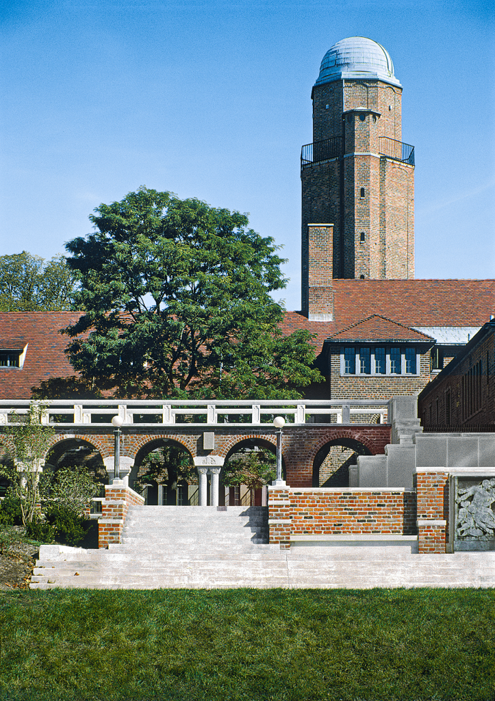 A tall brick observatory tower with a domed roof rises behind an arched brick walkway, mature trees, and a manicured lawn under a clear blue sky.