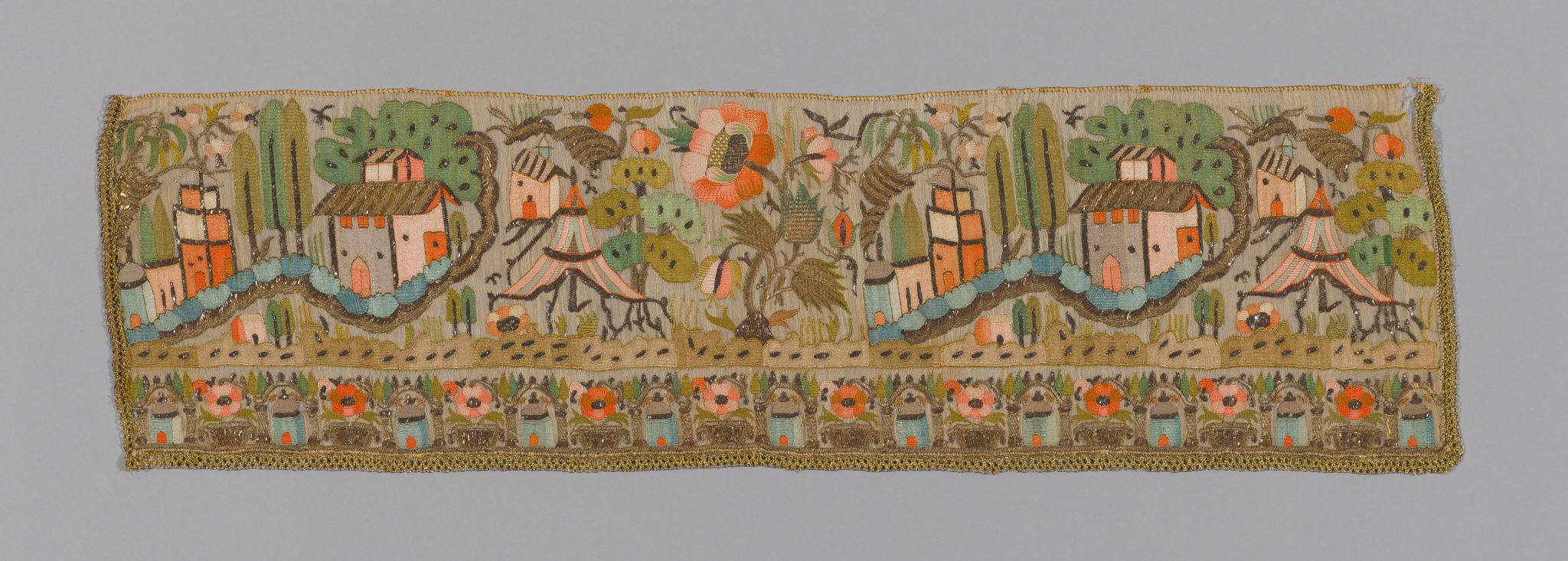 A rectangular textile panel embroidered with colorful thread depicting houses, trees, flowers, and a winding blue stream, bordered below by a row of flowers and edged with delicate lace trim.