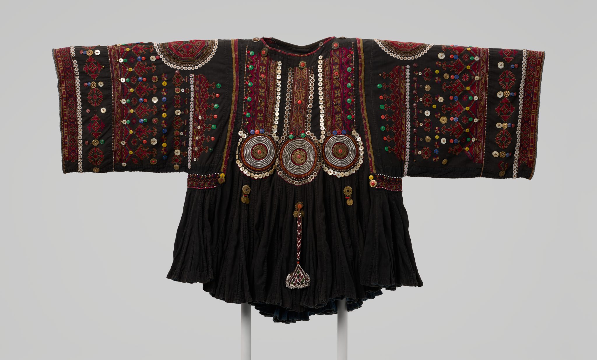 A richly decorated traditional garment with wide sleeves, featuring intricate embroidery, colorful sequins, beads, and circular metallic ornaments on dark fabric, displayed on a stand against a plain background.