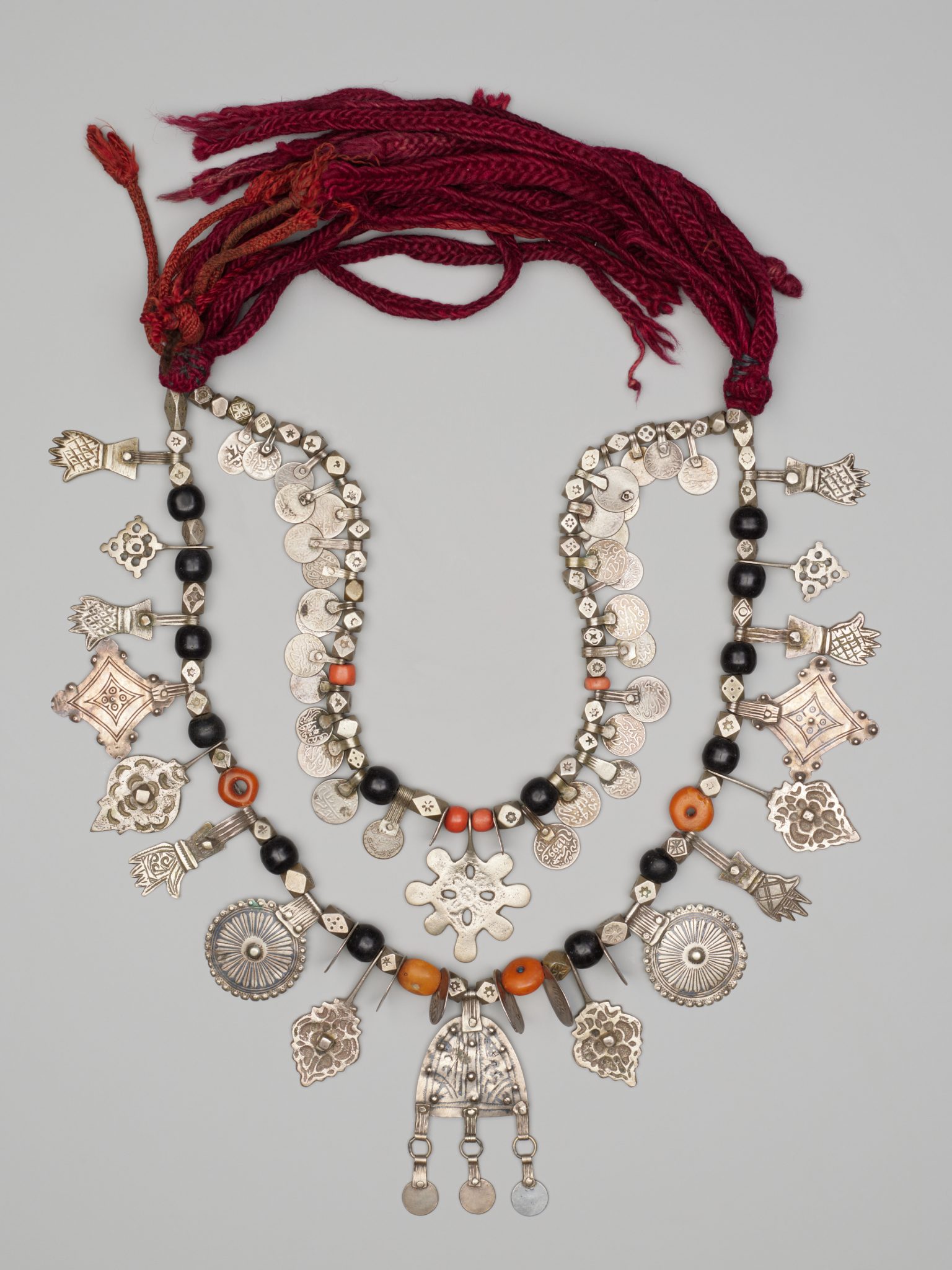 A two-tiered necklace with silver charms, coins, black and orange beads, and intricate metalwork, fastened with braided dark red cords. The charms are varied in shape, including floral, circular, and hand motifs.