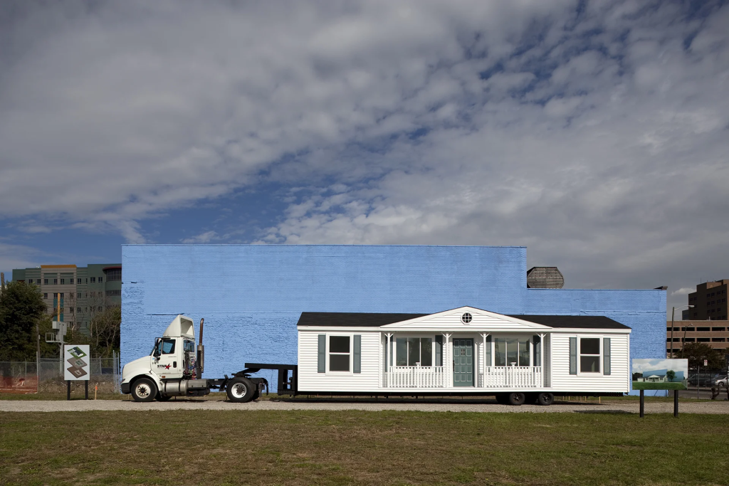 A white mobile home on a flatbed trailer is parked in front of a large blue-painted building under a partly cloudy sky. There are signs and grassy areas nearby.