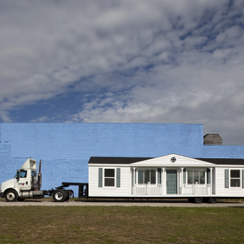 A white mobile home on a flatbed trailer is parked in front of a large blue-painted building under a partly cloudy sky. There are signs and grassy areas nearby.