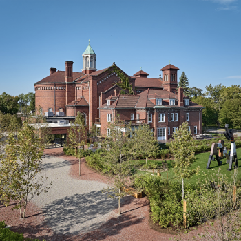A large red-brick building with a dome and tower, surrounded by trees and greenery, under a clear blue sky. Sculptures stand on the lawn near the building.