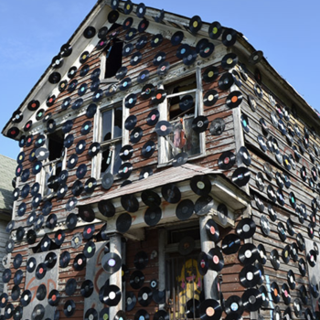 An old, weathered two-story house is decorated with numerous vinyl records attached to its exterior walls, under a clear blue sky.
