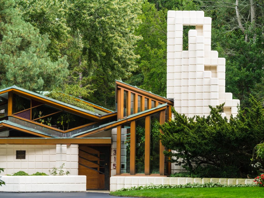 Modern house with geometric white blocks, angled wooden beams, large windows, and lush greenery surrounding it. The house number 315 is visible near the entrance.