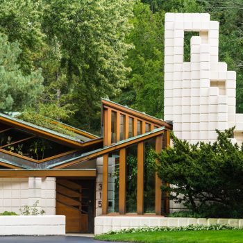 Modern house with geometric white blocks, angled wooden beams, large windows, and lush greenery surrounding it. The house number 315 is visible near the entrance.