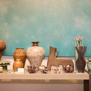 A variety of ceramic vases and pottery pieces are displayed on a table in front of a large teal artwork, showcasing different shapes, sizes, and textures.