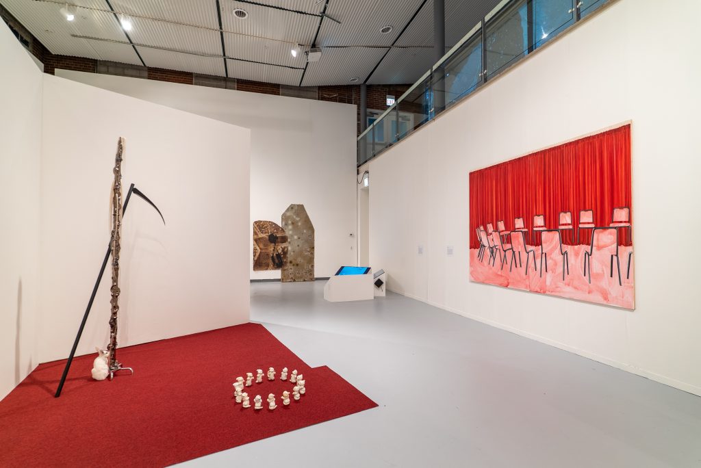 A modern art gallery with various installations: a scythe and skulls on a red carpet, a large pink painting of chairs and curtains, and assorted sculptural objects against white walls under high ceilings.