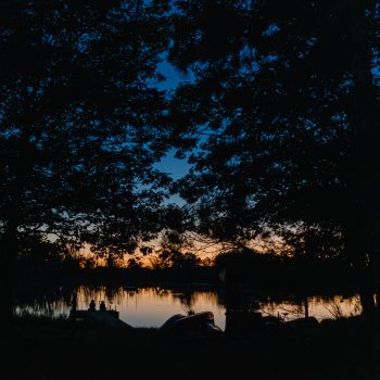 Silhouette of trees by a calm lake at sunset, with dark blue sky fading to orange near the horizon and faint reflections in the water.