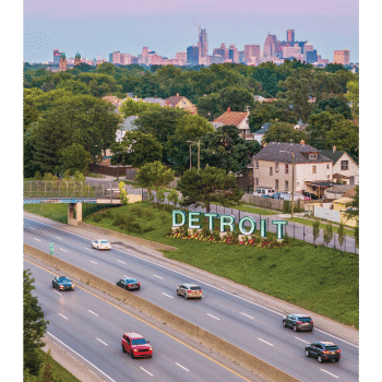 A highway runs past a large DETROIT sign with homes and trees nearby; the Detroit city skyline rises in the background under a partly cloudy sky.