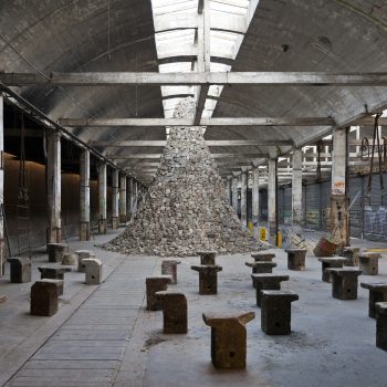 An industrial warehouse contains scattered stone stools and a large conical pile of bricks at the center, surrounded by graffiti-covered walls and exposed beams.