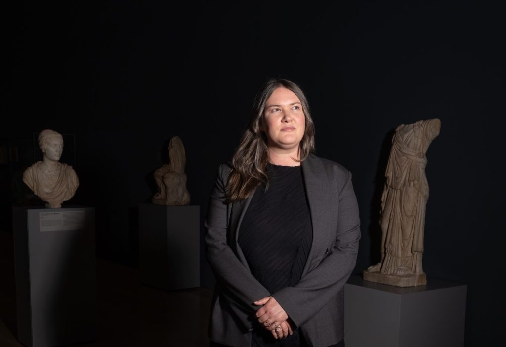 A woman in dark clothing and a gray blazer stands in a dimly lit room with classical statues on pedestals behind her, on a wooden floor.